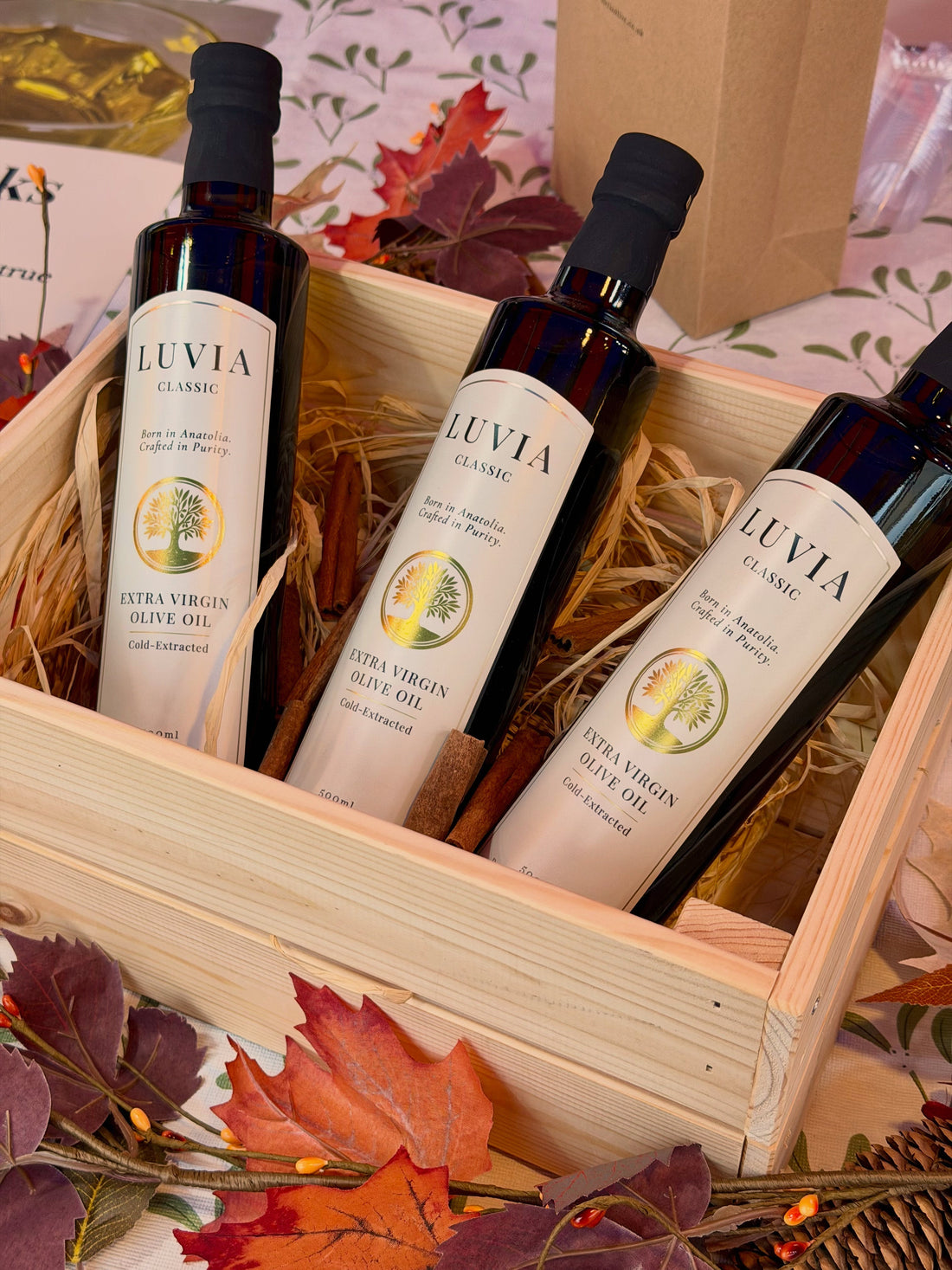 Three bottles of LUVIA Classic extra virgin olive oil in a wooden gift box surrounded by autumn leaves, representing premium Turkish olive oil available in the UK.
